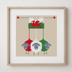 Op de afbeelding: Een ingelijst kruissteekkunstwerk met drie schapen in rood, wit en groen, elk met een andere kleur hoed. Boven de schapen is een groene en witte banner met een rode draak, het symbool van Wales. De achtergrond is lichtbruin.