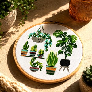 May include: A circular wooden embroidery hoop containing a white fabric panel with a cross-stitch pattern of various potted plants. The design features a hanging plant, a large leafy plant on a stand, and several smaller cacti and succulents in pots.