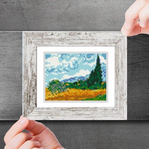 May include: A framed cross-stitch artwork depicting a landscape with a green tree, a field of yellow flowers, and a blue sky with white clouds.