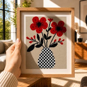 Puede incluir: Una obra de arte floral en punto de cruz enmarcada. La obra presenta flores rojas con centros negros y hojas en un jarrón a cuadros. El marco es de madera marrón claro.