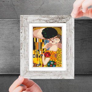 May include: A framed cross-stitch of Gustav Klimt's "The Kiss." The artwork depicts a couple embracing, in a mosaic of gold, yellow, and colourful squares. The frame is distressed white wood.