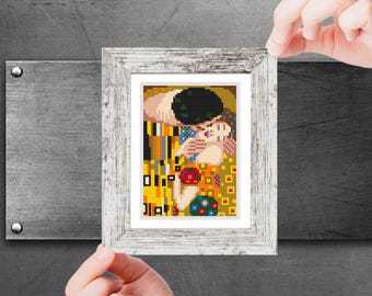 Pode incluir: Uma bordado em ponto cruz emoldurado de "O Beijo" de Gustav Klimt. A obra de arte apresenta um casal se abraçando, renderizado em um mosaico de quadrados dourados, amarelos e coloridos. A moldura é de madeira branca desgastada.