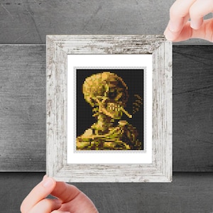 May include: A framed cross-stitch artwork depicting a yellow skull with a cigarette in its mouth. The skull is wearing a hat and has a detailed, pixelated design.