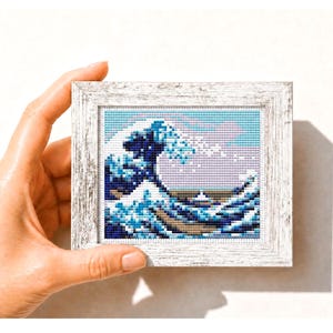 Könnte beinhalten: Ein gerahmtes Kreuzstichbild von Hokusais "Die große Welle vor Kanagawa". Das Kunstwerk verwendet Blau- und Weißtöne, mit einem hellblauen Himmel und einem weißen Holzrahmen. Das Bild wird von einer Hand gehalten.
