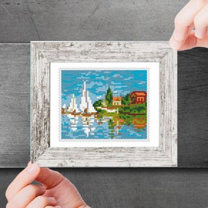 May include: A framed cross-stitch artwork depicting a small village with white sailboats on a blue body of water. The scene includes a small house, trees, and a blue sky with white clouds.