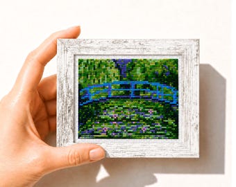 Op de afbeelding: Een ingelijst kruissteekkunstwerk met een blauwe brug over een tuin met groene bladeren en paarse bloemen. De witte lijst heeft een verweerde afwerking. Het kunstwerk wordt door een hand vastgehouden.