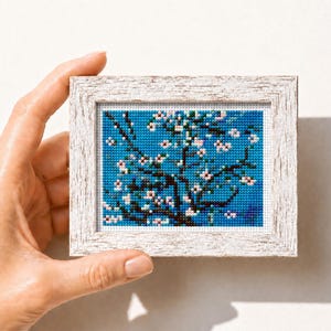 Könnte beinhalten: Ein gerahmtes Kreuzstich-Kunstwerk mit einem Baum mit weißen und rosa Blüten vor blauem Hintergrund. Der Rahmen ist weiß mit einem Used-Look. Das Kunstwerk wird von einer Hand gehalten.