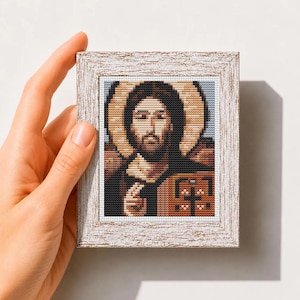 Puede incluir: Un patrón de punto de cruz con un icono religioso de una figura con halo, cabello oscuro y barba, sosteniendo un libro con una cruz. La obra está en un marco de madera blanca desgastada. El texto dice Mini Art Cross Stitch Pattern PDF.