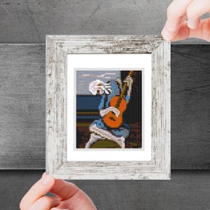 May include: A framed cross-stitch artwork depicting a person playing a guitar. The person is wearing a blue shirt and the guitar is orange. The background is a blue and white sky.