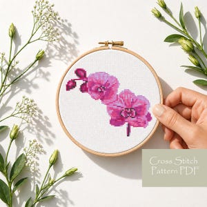 Puede incluir: Un patrón de punto de cruz de una flor de orquídea rosa en un bastidor de bordado de madera. El texto "Cross Stitch Pattern PDF" está en un rectángulo verde claro. El fondo es blanco con follaje verde. El bastidor mide unos 15 cm de diámetro.