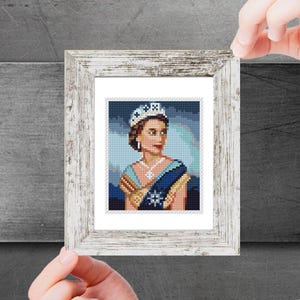 May include: A framed cross-stitch portrait of a woman wearing a crown, necklace, and blue sash. The portrait is in a white distressed wooden frame. The background is a gradient of blue and green.
