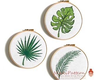 Op de afbeelding: Drie ronde borduurringen met kruissteekontwerpen van tropische bladeren. De bladeren zijn in verschillende groentinten, waaronder een gatenplant, een palmblad en een waaierpalm. De ringen hebben een natuurlijke houten afwerking en zijn op een witte achtergrond gemonteerd.