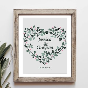 Könnte beinhalten: Gerahmtes Kreuzstich-Kunstwerk mit einem herzförmigen Kranz aus grünen Blättern und rosa Beeren. Die Namen "Jessica & Greyson" sind zentriert, darunter das Datum "10.09.2025". Der Rahmen ist hellbraun. Das Kunstwerk ist in einem Holzrahmen.