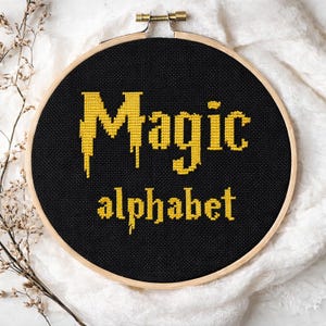 Puede incluir: Un bastidor de punto de cruz negro con las palabras "Magic alphabet" en amarillo. El texto está en una fuente estilizada. El bastidor se encuentra sobre un fondo de tela blanca. El aro de madera está asegurado con un cierre de latón.