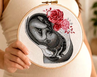 Op de afbeelding: Een kruissteek borduurring met een grijswaarden afbeelding van een foetus, met rode en roze rozen en bloemaccenten. De borduursel zit in een houten ring.