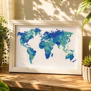 May include: A framed cross-stitch of a world map, featuring continents in shades of blue and green against a white background. The artwork is displayed in a white frame and rests on a wooden surface. The piece is a decorative item.