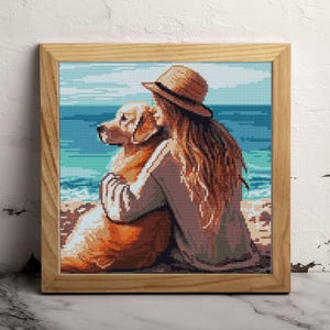 May include: A framed cross-stitch artwork depicts a person hugging a golden retriever on a beach. The person wears a straw hat and has long, flowing auburn hair. The background shows a blue ocean and sky.