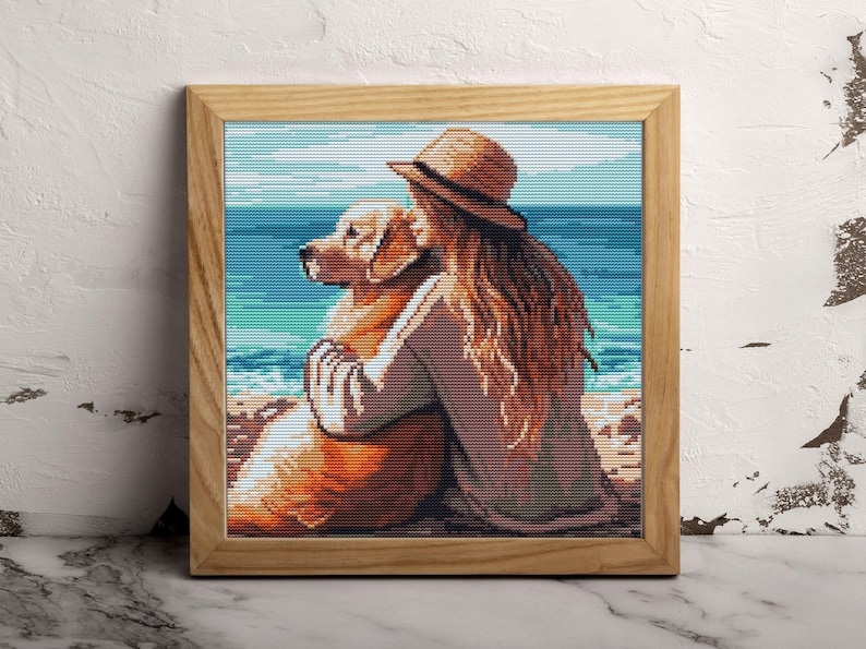 May include: A framed cross-stitch artwork depicts a person hugging a golden retriever on a beach. The person wears a straw hat and has long, flowing auburn hair. The background shows a blue ocean and sky.