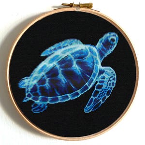May include: A cross-stitch embroidery of a sea turtle in shades of blue against a black background. The turtle is centered within a round wooden hoop, showcasing the detailed needlework. The hoop is approximately 8 inches in diameter.