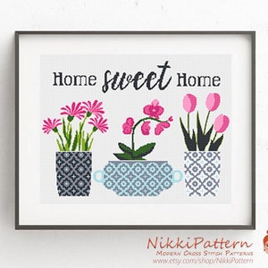 May include: Cross stitch pattern featuring three potted plants with the text "Home sweet Home". The plants are a pink orchid, pink tulips, and pink flowers with green leaves. The pots are gray and white with geometric patterns.