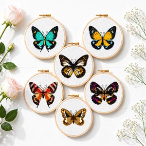 May include: Six embroidered butterfly designs in wooden hoops. Each butterfly has a unique colour scheme, including teal, yellow, orange, and purple, with black detailing. The hoops are arranged on a white surface, with pink roses and white flowers.