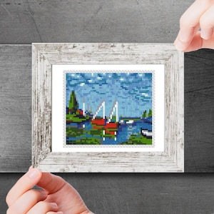 Könnte beinhalten: Ein gerahmtes Kreuzstich-Kunstwerk, das eine Seenszene mit Segelbooten und einem roten Haus in der Ferne zeigt. Der Himmel ist blau mit weißen Wolken. Das Kunstwerk ist in einem weißen Holzrahmen gerahmt.