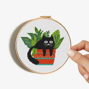 May include: Cross stitch pattern of a black cat sitting in a red and green pot with green leaves. The cat has white eyes and a black nose.