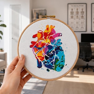 May include: A colorful cross-stitch embroidery of a human heart, held in a wooden hoop. The heart design features vibrant hues of red, orange, yellow, pink, purple, and blue. The artwork is displayed on a white fabric.