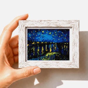 Puede incluir: Una pequeña obra de arte en punto de cruz enmarcada que representa un paisaje urbano nocturno. La escena presenta un cielo azul oscuro con estrellas amarillas y una silueta de edificios con luces amarillas. El marco es blanco con un acabado envejecido.