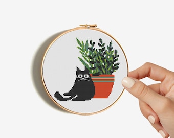 Op de afbeelding: Kruissteekborduurwerk in een houten hoepel met een zwarte kat en een potplant. De kat zit naast een terracotta pot met groen gebladerte. De hoepel wordt door een hand tegen een witte achtergrond gehouden.