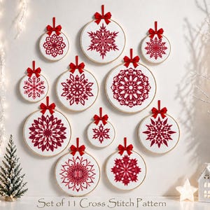 May include: A set of 11 cross-stitch patterns featuring red snowflake designs on white fabric, framed in embroidery hoops. Each hoop is adorned with a red ribbon for hanging. The text "Set of 11 Cross Stitch Pattern" is visible at the bottom.
