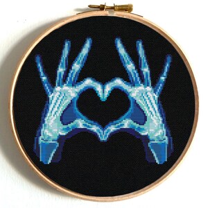 Könnte beinhalten: Kreuzstich-Stickerei in einem Holzreifen mit schwarzem Hintergrund und einem blau-weißen Bild von Händen, die ein Herz formen. Die Hände sind als Röntgenaufnahme dargestellt, die das Skelett zeigt.