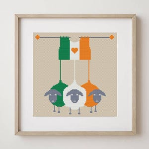 May include: Framed cross-stitch artwork. Three sheep in green, white, and orange hang below flags of the same colours, with a heart on the white flag. The artwork is in a light wood frame, ideal for home decor.