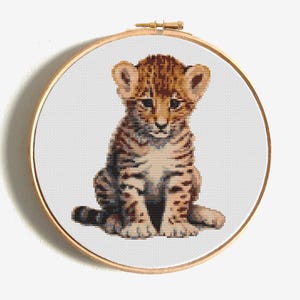 Puede incluir: Un bordado de punto de cruz de un cachorro de tigre bebé, con tonos marrones, beige y blanco. El cachorro está sentado, con orejas grandes y una mirada concentrada. El bordado está en un aro de madera redondo.