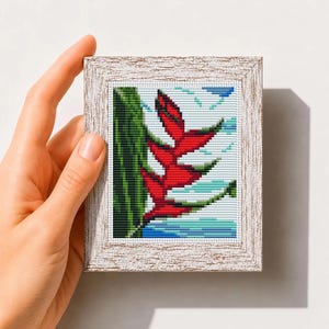 May include: A cross stitch pattern featuring a vibrant red heliconia flower against a green leaf and blue sky background, framed in a light-colored wood frame. The text "Mini Art Cross Stitch Pattern PDF" is printed at the bottom of the image.