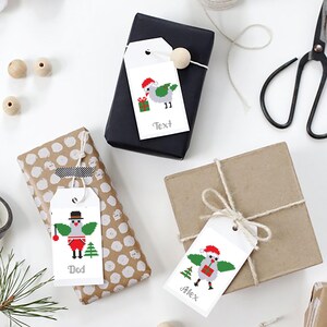 Puede incluir: Tres regalos envueltos con etiquetas decorativas. Las etiquetas presentan dise&ntilde;os de p&aacute;jaros navide&ntilde;os pixelados en rojo, verde y blanco. Una etiqueta dice "Text", otra "Dad" y la tercera "Alex". Los regalos est&aacute;n envueltos en papel negro, marr&oacute;n y de lunares.