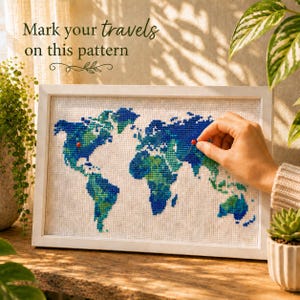 May include: A framed cross-stitch world map in shades of blue and green, with red pins marking locations. The white frame surrounds the map, which is displayed on a wooden surface. Text at the top reads "Mark your travels on this pattern."