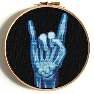 Könnte beinhalten: Ein Kreuzstich-Design einer Hand, die die Rock-and-Roll-Geste macht, in Blau und Weiß vor einem schwarzen Hintergrund. Das Design ist in einem runden Holz-Stickrahmen mit Messingverschluss gerahmt.