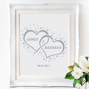 Op de afbeelding: Een ingelijst kruissteekkunstwerk met twee grijze, in elkaar grijpende harten met de namen "JAMES" en "BARBARA". De datum "08.06.2021" staat eronder. Het kunstwerk zit in een witte, sierlijke lijst.