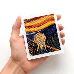 May include: A small, rectangular cross-stitch of Edvard Munch's "The Scream" held in a hand. The artwork features a figure with a screaming expression against a vibrant, swirling sky of orange, yellow, and blue. The cross-stitch is on a white background.