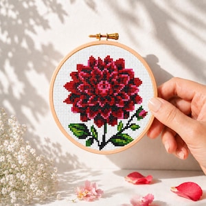 May include: A circular wooden embroidery hoop containing a cross-stitch pattern of a large, dark red dahlia flower with a bright pink center, green leaves, and a small bud, set against a white fabric background with a metallic tension screw.