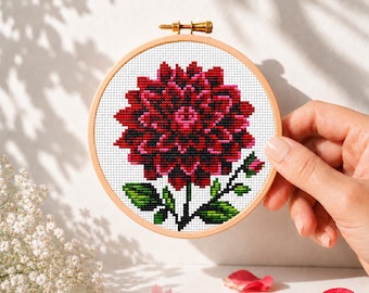 May include: A circular wooden embroidery hoop containing a cross-stitch pattern of a large, dark red dahlia flower with a bright pink center, green leaves, and a small bud, set against a white fabric background with a metallic tension screw.
