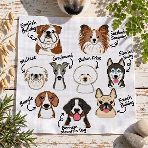 May include: A white fabric square featuring cross-stitch portraits of nine dog breeds: English Bulldog, Shetland Sheepdog, Maltese, Greyhound, Bichon Frise, Siberian Husky, Beagle, Bernese Mountain Dog, and French Bulldog, each with its name labelled.