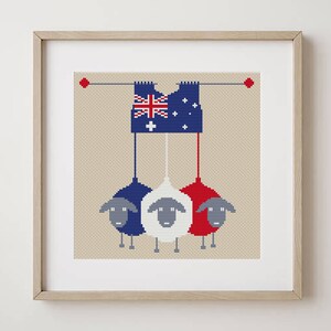 May include: Framed cross-stitch art featuring three sheep in red, white, and blue, hanging from a banner with the Australian flag. The artwork is set against a neutral background and framed in a light wood.