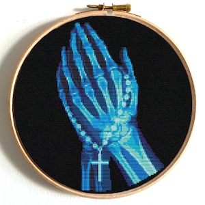 May include: A cross-stitch embroidery in a wooden hoop. The design features a pair of hands in a praying position, rendered in a blue skeletal style against a black background. A rosary with a cross hangs between the hands.