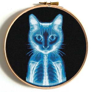 May include: A blue and white cross-stitch embroidery of a cat's skeleton, with a black background. The cat is sitting upright with its head tilted slightly to the side. The embroidery is framed in a wooden hoop.