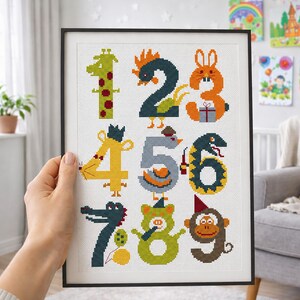 Puede incluir: Obra de arte en punto de cruz enmarcada con los números del 1 al 9, cada uno diseñado como un animal diferente. Los números son de varios colores, incluyendo verde, naranja, amarillo y marrón. La obra está en un marco negro.