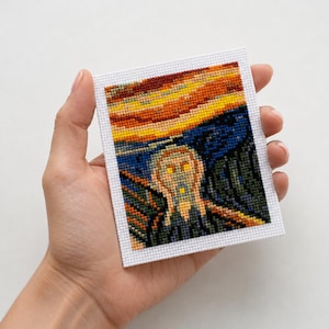 May include: A cross-stitch of Edvard Munch's "The Scream" artwork. The piece features a figure with an open mouth against a vibrant sunset sky. The artwork is framed in white and held in a hand.