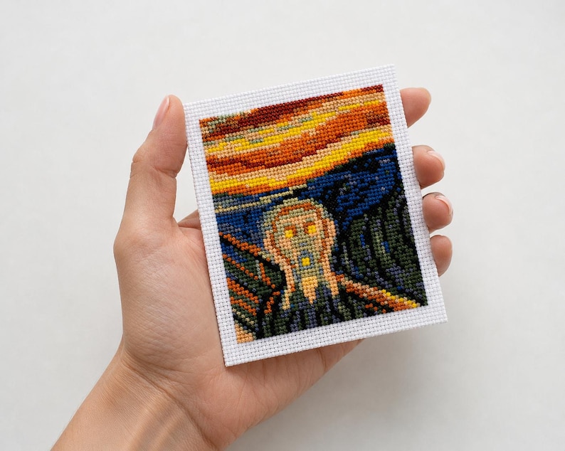 May include: A cross-stitch of Edvard Munch's "The Scream" artwork. The piece features a figure with an open mouth against a vibrant sunset sky. The artwork is framed in white and held in a hand.