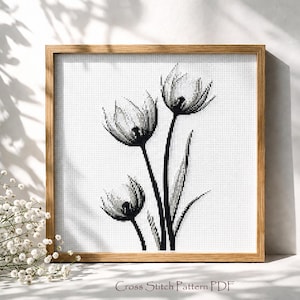 Peut inclure: Un motif de point de croix encadré représentant trois fleurs stylisées en noir et blanc. L'œuvre est placée sur un fond blanc et encadrée dans un cadre en bois brun clair. Le texte "Cross Stitch Pattern PDF" est en bas.